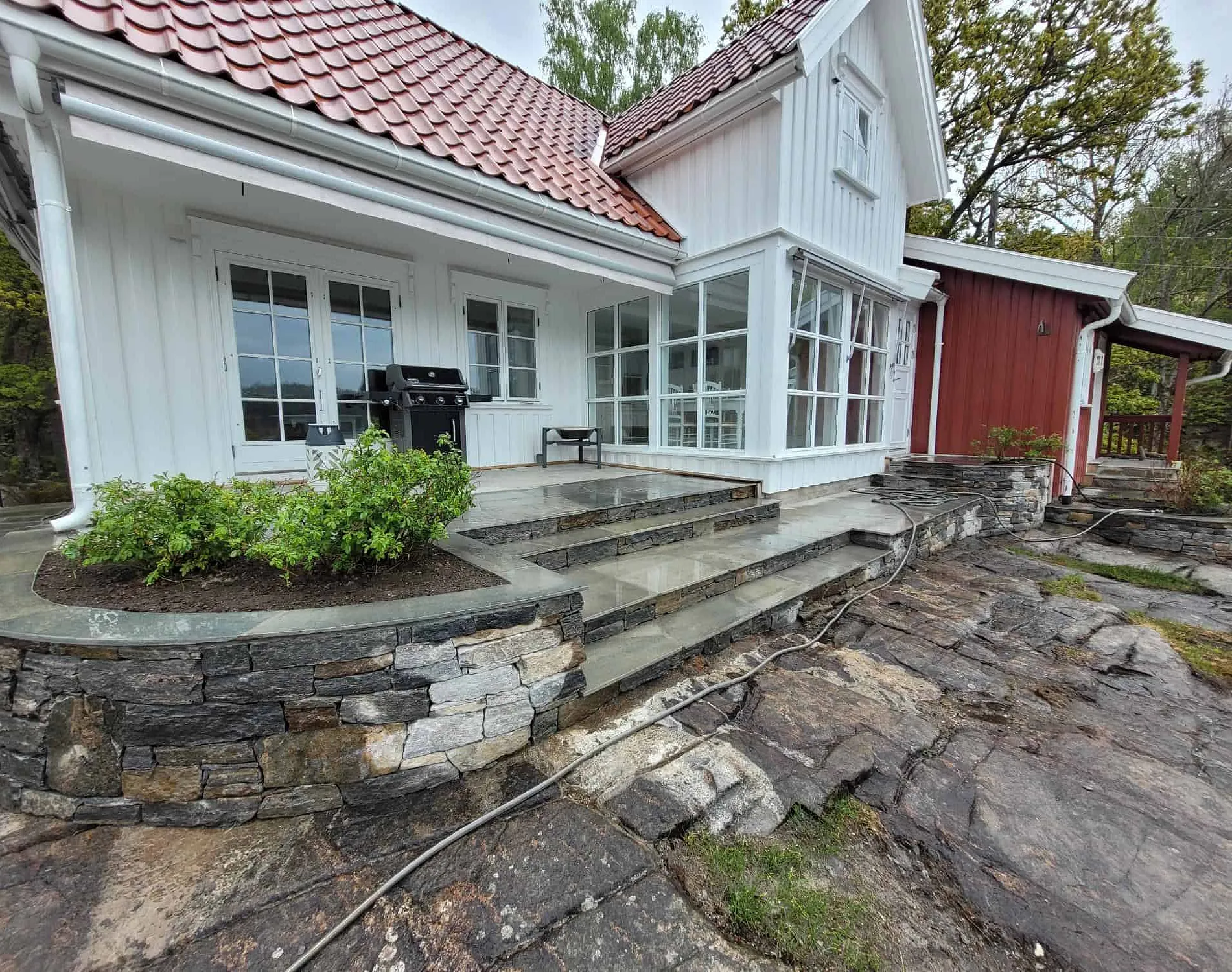 Steinlagt terrasse med blomsterbed ved et hvitt hus med rødt anneks