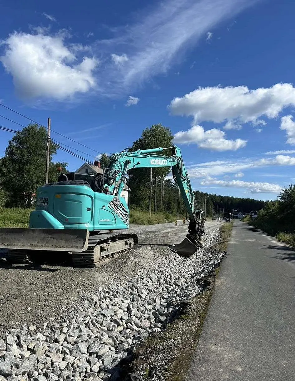 Gravemaskin legger pukk langs en grusvei under blå himmel