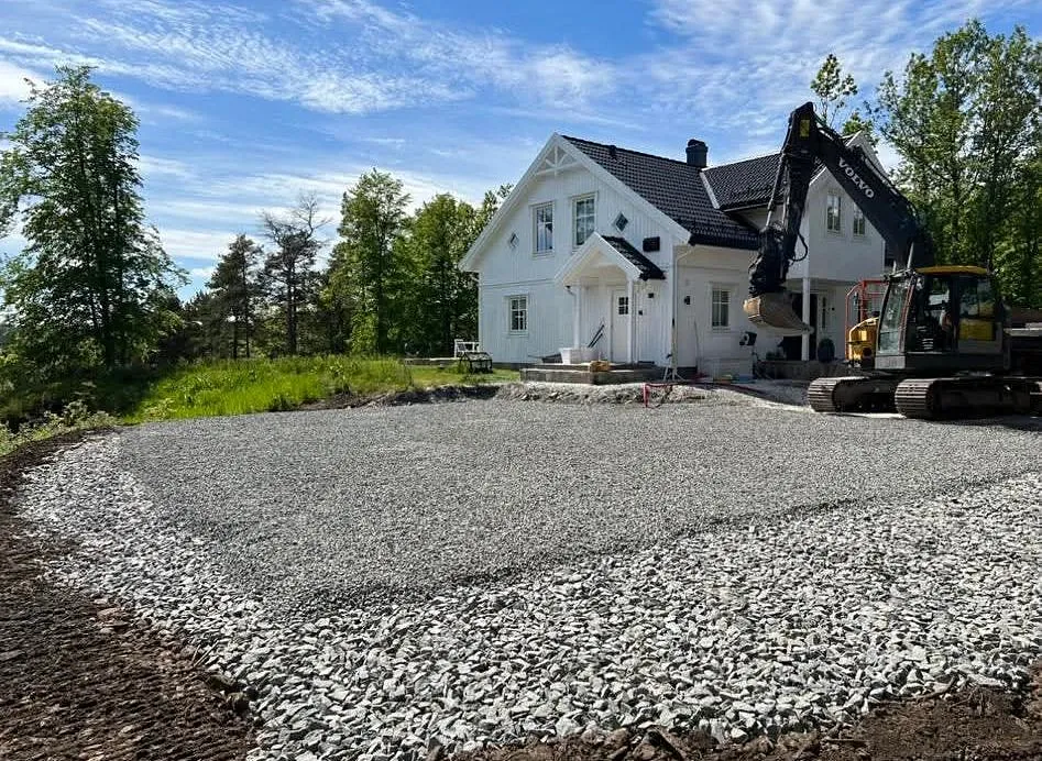 Gruslagt gårdsplass under arbeid ved et hvitt hus med gravemaskin