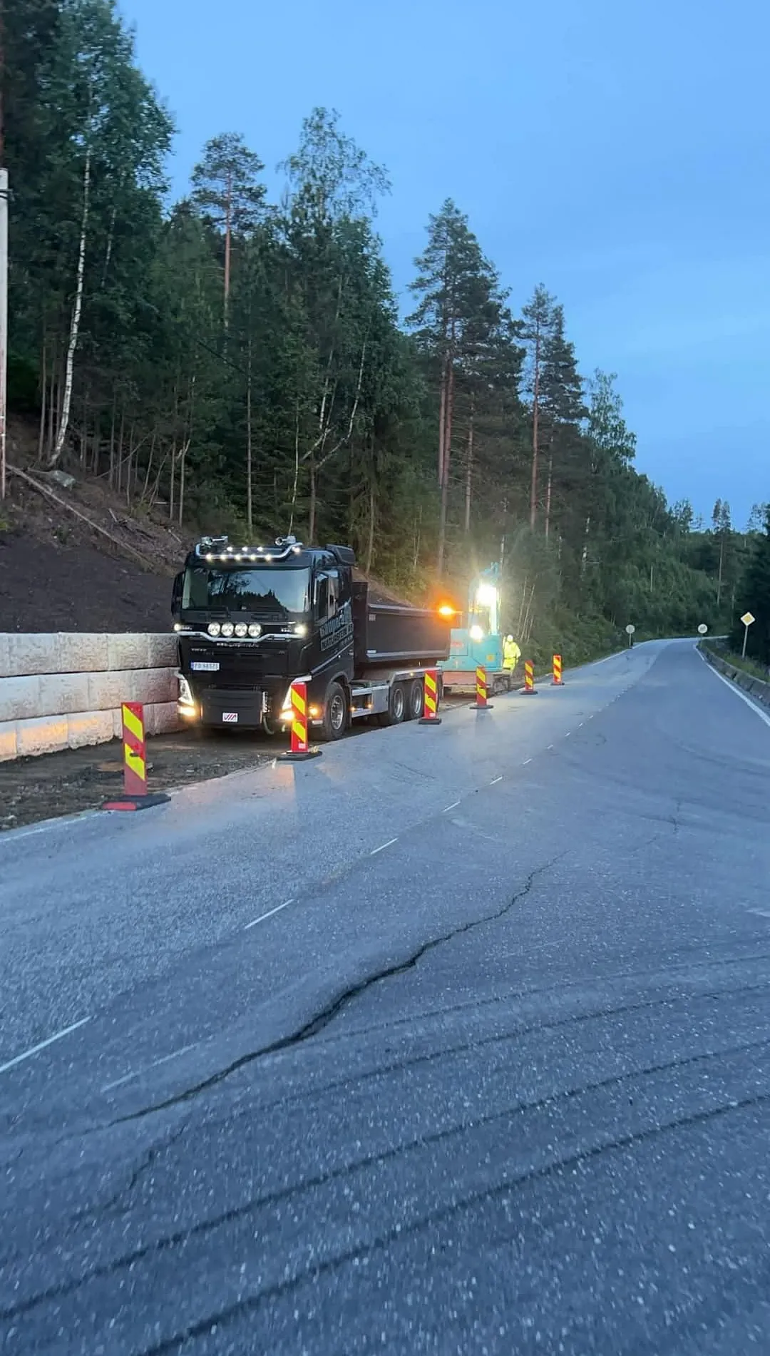 Lastebil og gravemaskin arbeider langs en opplyst vei i skogsområde